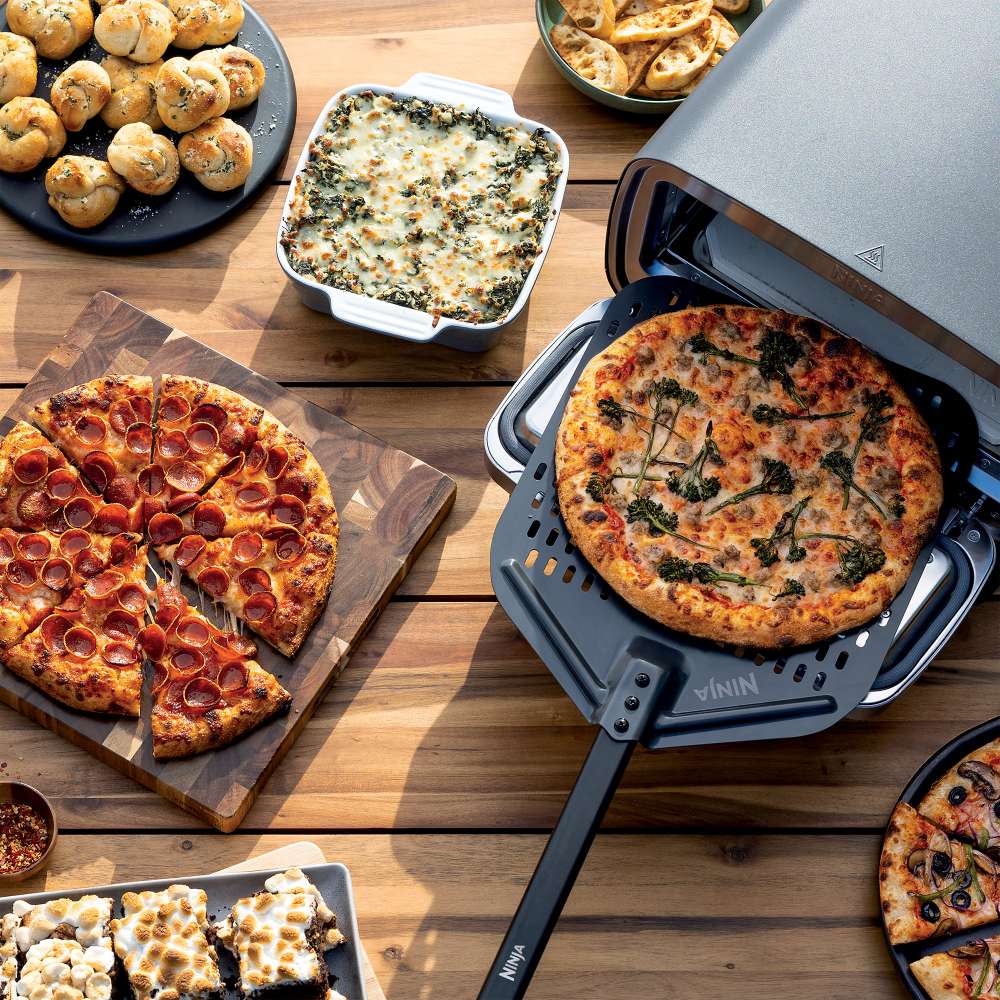 Pizza, baked pasta, rolls, and dessert surround the Ninja Artisan 5-in-1 Portable Electric Pizza & Outdoor Oven on an outdoor table
