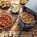 Pizza, baked pasta, rolls, and dessert surround the Ninja Artisan 5-in-1 Portable Electric Pizza & Outdoor Oven on an outdoor table