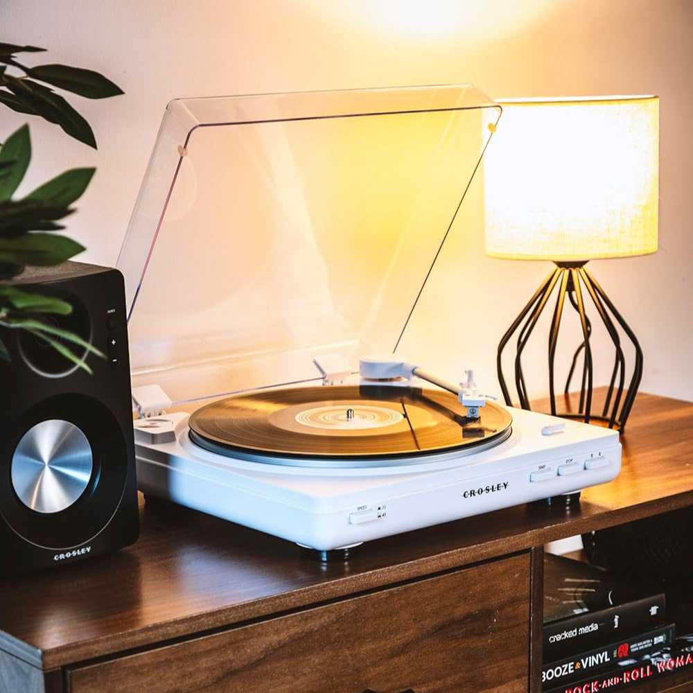 Playing a vinyl record, the Crosley T400D Component Bluetooth Turntable - White adds charm to a wooden console, softly lit by a table lamp