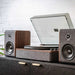 Positioned against a white brick wall, the Crosley Nocturne Record Player With Speakers - Charcoal features a minimalist design with matching speakers