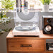 Positioned next to a speaker, the Crosley T400D Component Bluetooth Turntable - White spins a vinyl record with books and plants in the background