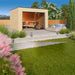 A landscaped backyard scene features the GardenHouse24 USA Q Bic ALU 44A Garden Room furnished with cozy seating and surrounded by greenery and a mountain backdrop