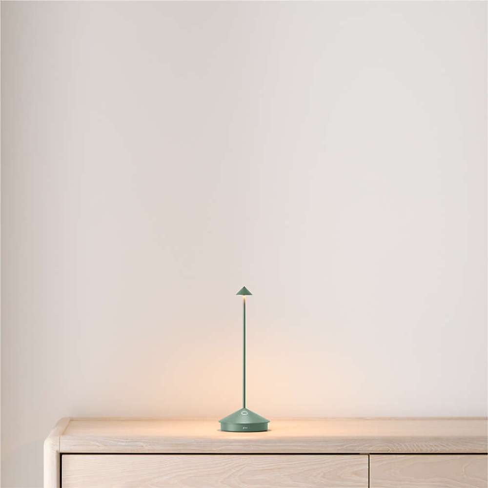 Sage Crate & Barrel Pina Pro Metal Table Lamp by Zafferano America glows warmly on a light wood console against a neutral wall