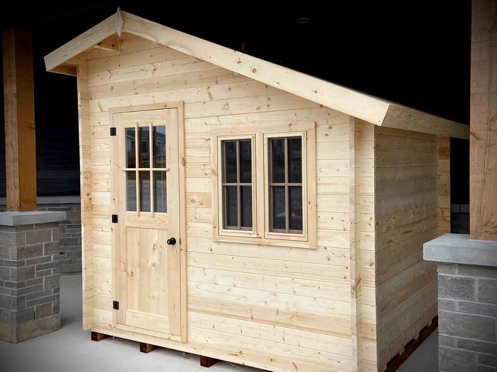Sawmill Structures Bluewater Den 67 Garden Room, a small wooden cabin with a front door and twin-paned windows under a slanted roof