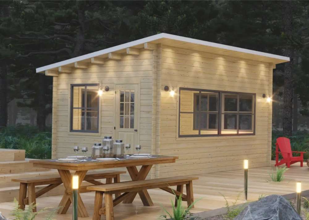 Sawmill Structures Eze Breeze Sunroom Cabin glows warmly at night, with a picnic table, deck lighting, and tall trees