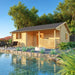 Wooden Sunbeam Lakehouse 70mm cabin with a covered front porch, patio furniture, fire feature, and lush landscaping beside a reflective pond.