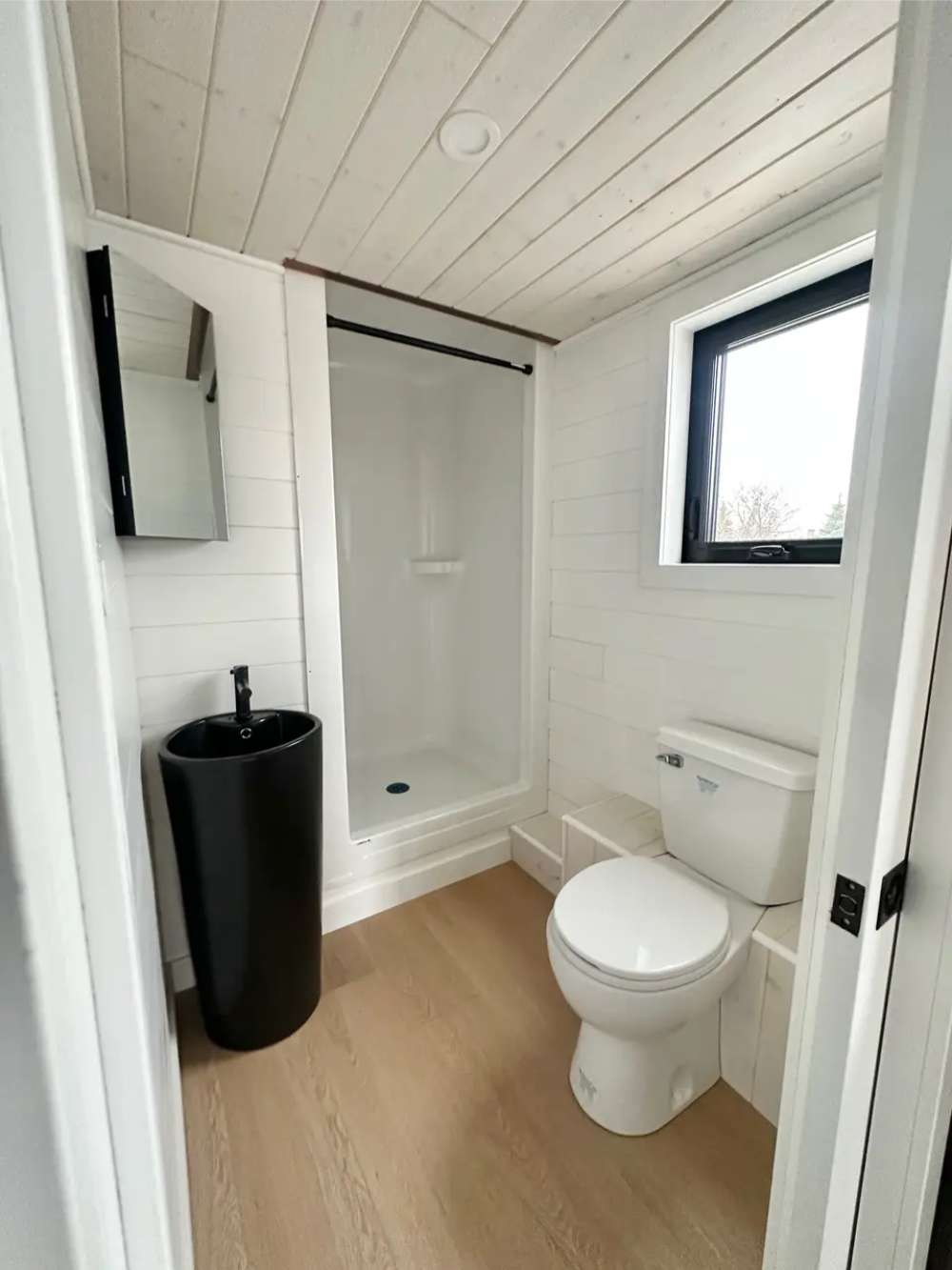 Sawmill Structures Wanderlove Tiny Home bathroom includes a white toilet, tall black sink, walk-in shower, and a window for natural light