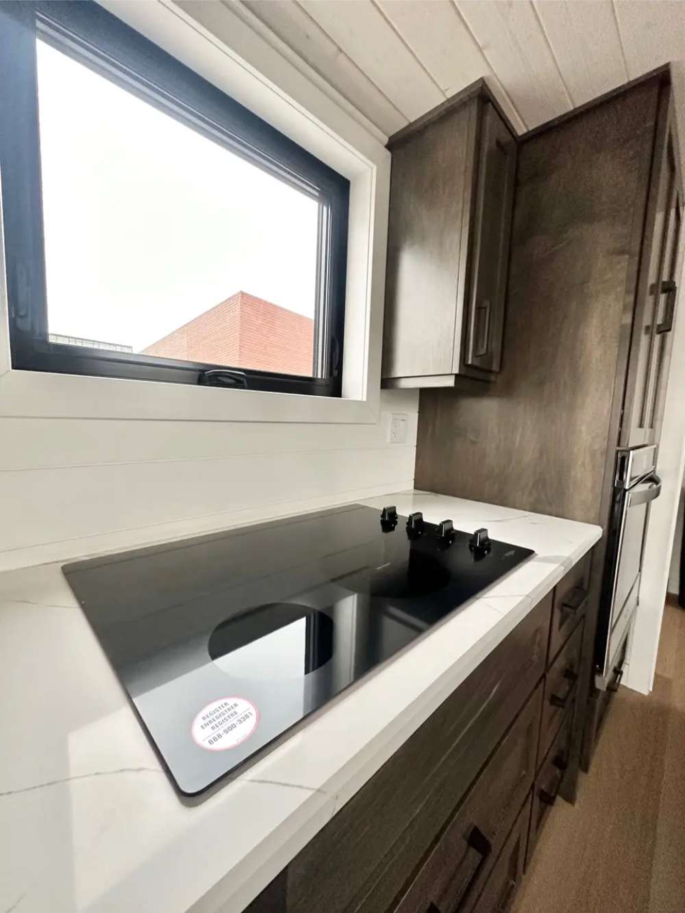 Sawmill Structures Wanderlove Tiny Home features a sleek black glass cooktop built into a white marble-patterned counter beneath a large window