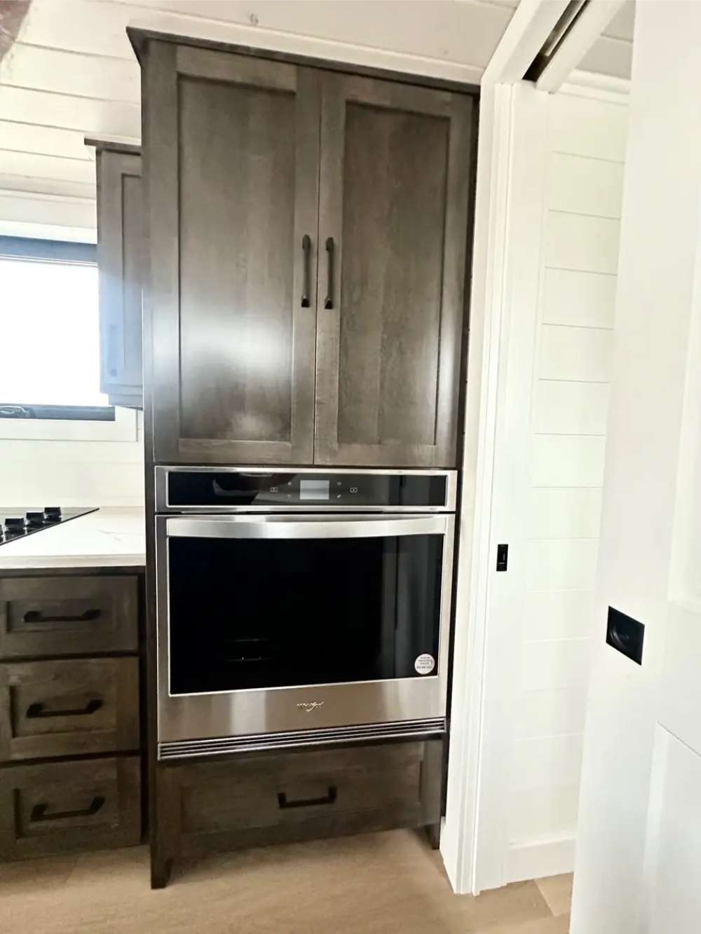 Sawmill Structures Wanderlove Tiny Home includes a stainless steel wall oven built into dark wood cabinetry next to a white sliding door