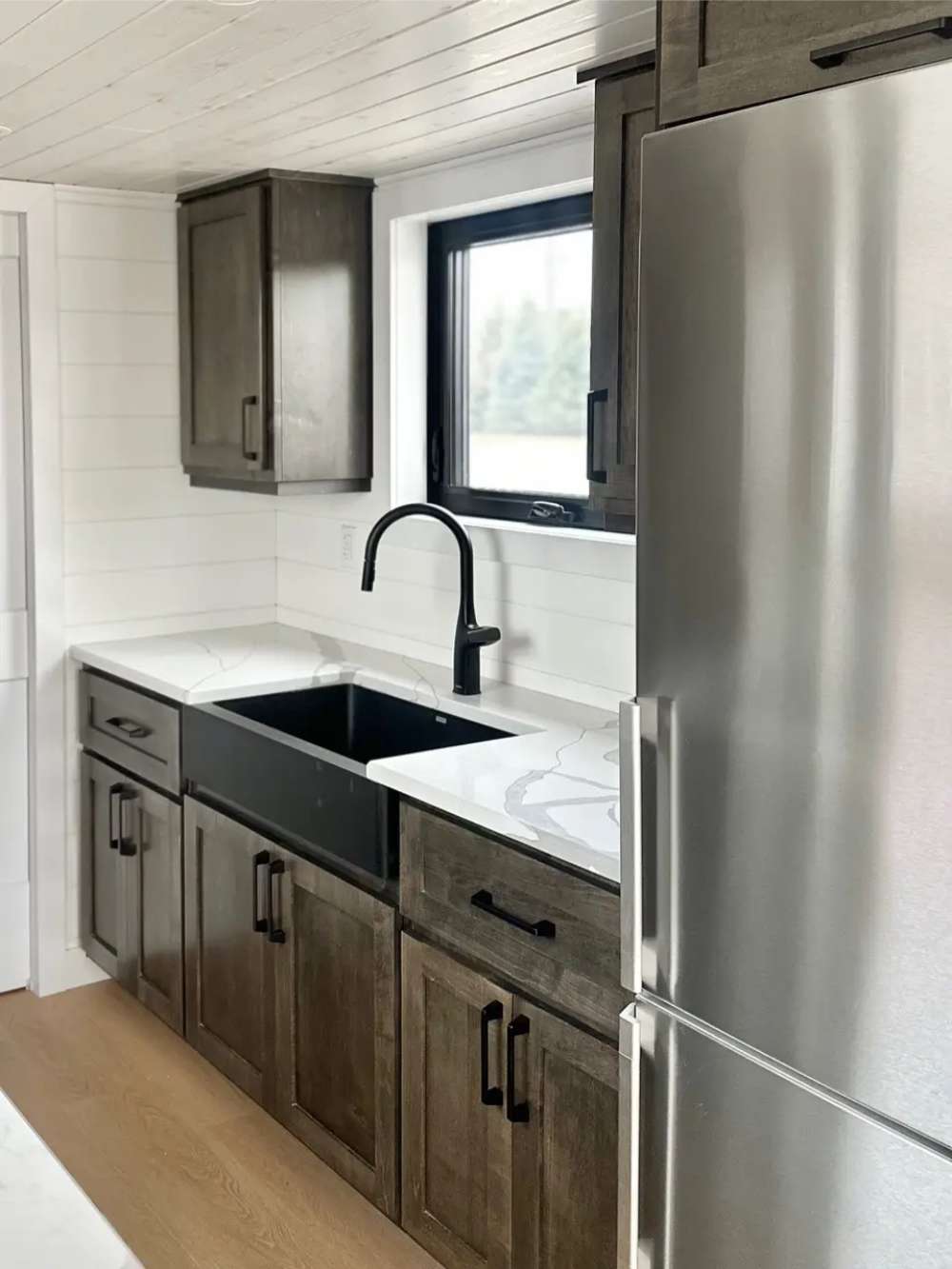 Sawmill Structures Wanderlove Tiny Home kitchen shows a modern black sink, marble-style counter, stainless fridge, and matching dark cabinets