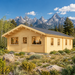 Rectangular light wood cabin with a gabled roof and multiple windows, set in a field with mountains in the background.