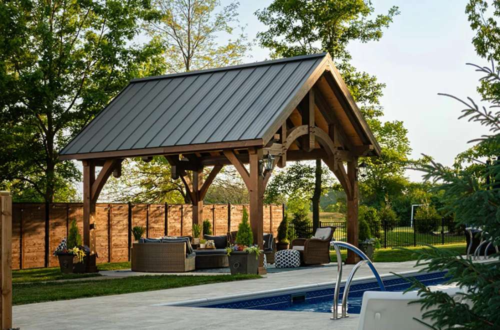 Set against lush greenery, the Brookside Timber Frame Alpine Lodge Pavilion blends rustic elegance with modern outdoor living in a tranquil backyard setting