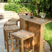 Set of several Westminster Teak Somerset Backless Bar Stool pieces is arranged around a wooden outdoor bar on a brick patio with greenery and flowers behind