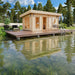 Set in a lush garden, the GardenHouse24 USA Sauna Sahib 70 Garden Room features large windows, warm lighting, and a round wooden wall detail, creating a peaceful retreat