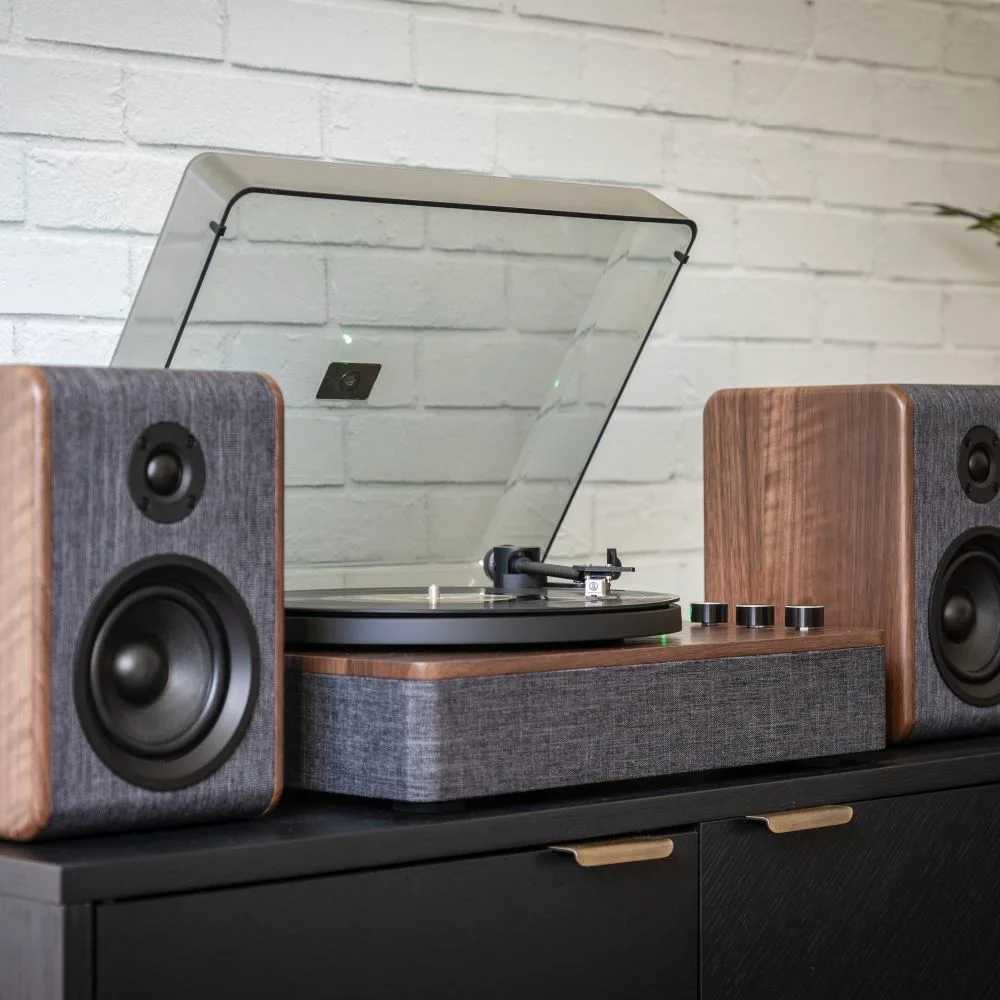 Set against a white brick wall, the Crosley Nocturne Record Player With Speakers - Charcoal plays a vinyl with its two speakers symmetrically placed