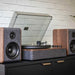 Set against a white brick wall, the Crosley Nocturne Record Player With Speakers - Charcoal plays a vinyl with its two speakers symmetrically placed