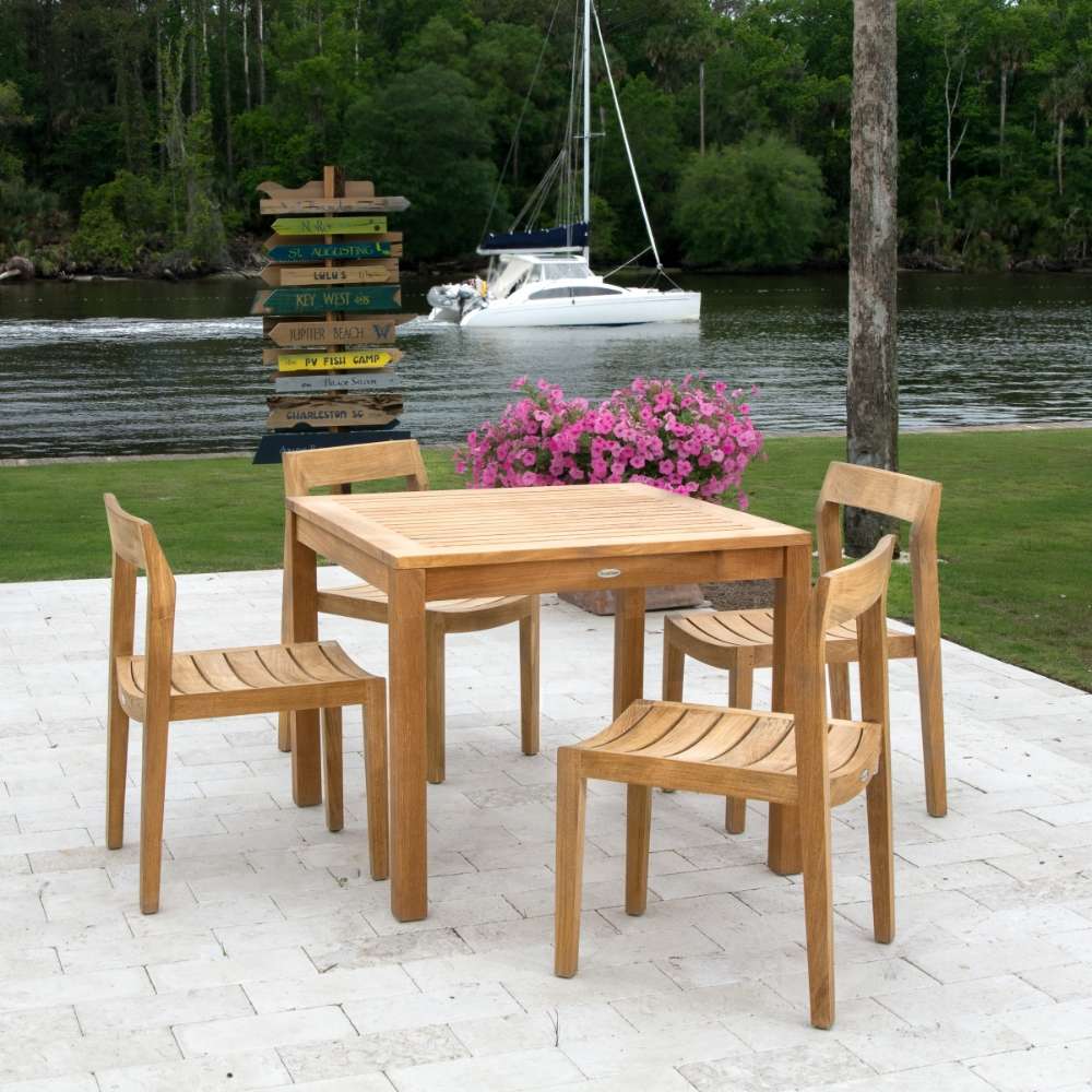 Several Westminster Teak Horizon Teak Side Chair pieces surround a wooden table on a patio near a lake, with a boat visible in the background