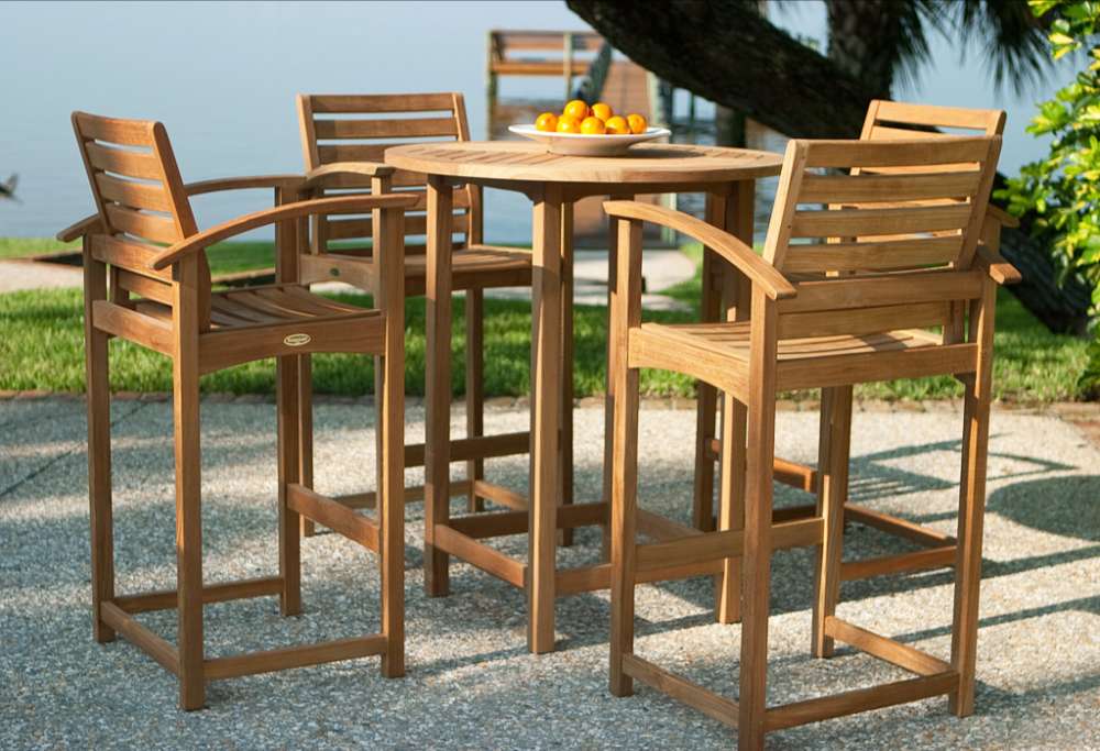 Several Westminster Teak Somerset Teak Bar Stools are arranged around a round outdoor bar table with a bowl of oranges, set on a patio overlooking greenery and water