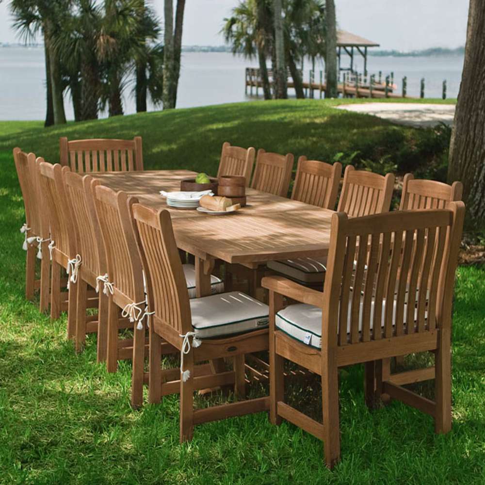 Several Westminster Teak Veranda Teak Armchairs are arranged around a large table on grass near palm trees and a lake, forming a spacious outdoor dining area
