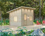 Single-room wooden cabin with a slanted roof sits on a wooden deck surrounded by tall forest trees—this is the Sawmill Structures Multi Cube Versa 100 Garden Room