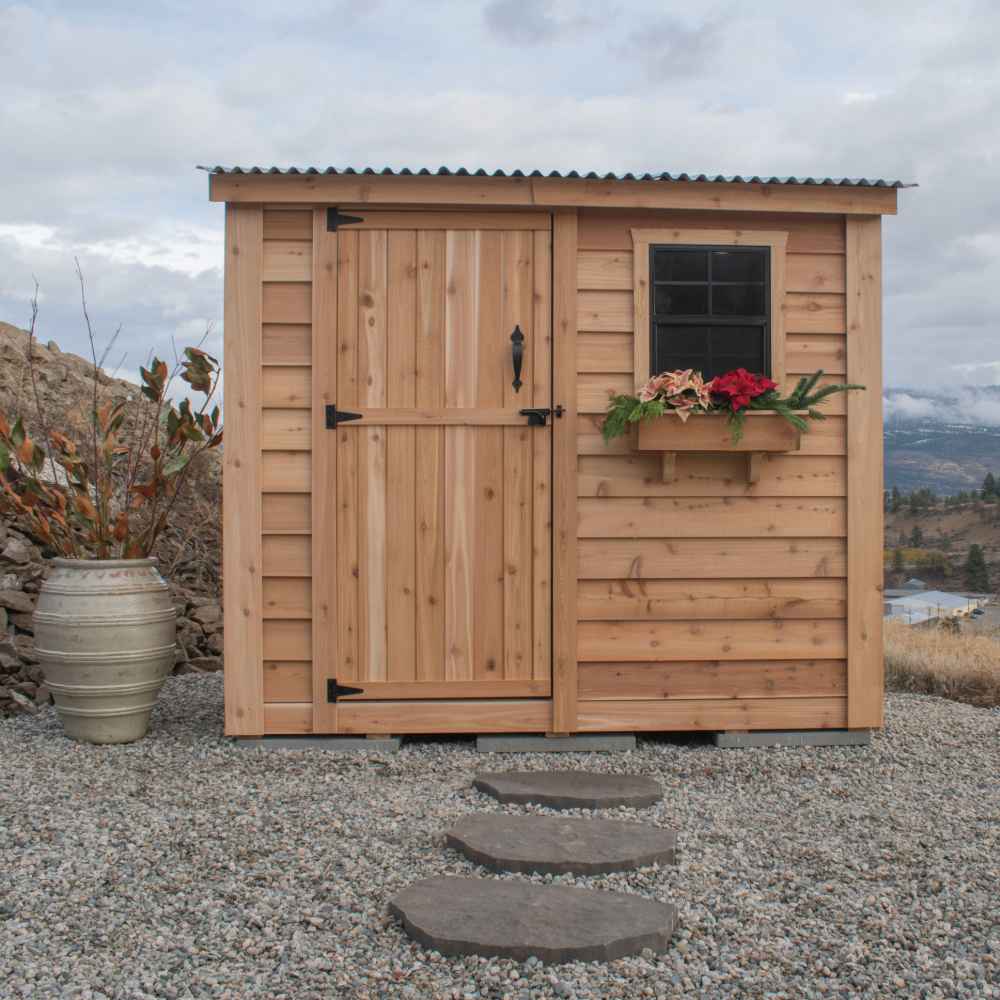 Single Door Metal Roof Outdoor Living Today 8×4 SpaceSaver Lean-To Cedar Shed stands on gravel with a vertical cedar door, metal roof, and a window decorated with festive flowers