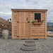 Single Door Metal Roof Outdoor Living Today 8×4 SpaceSaver Lean-To Cedar Shed stands on gravel with a vertical cedar door, metal roof, and a window decorated with festive flowers