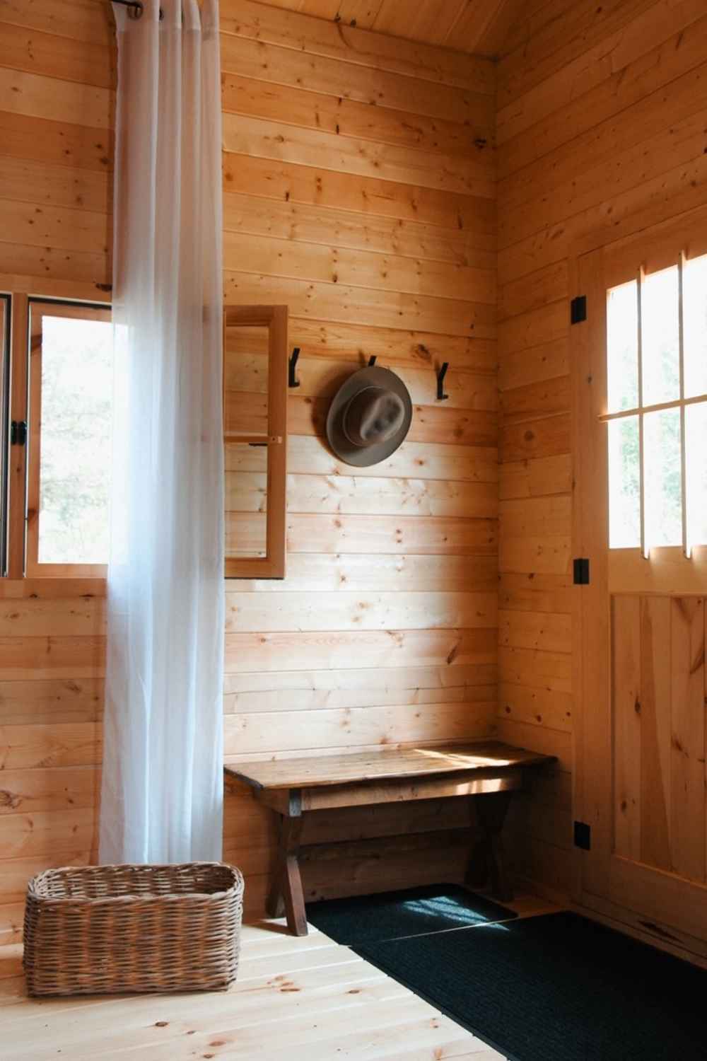 Small bench, hat rack, wicker basket, and tall curtains decorate the bright wooden entryway of the Sawmill Structures BlueWater Loft 160 38mm Garden Room
