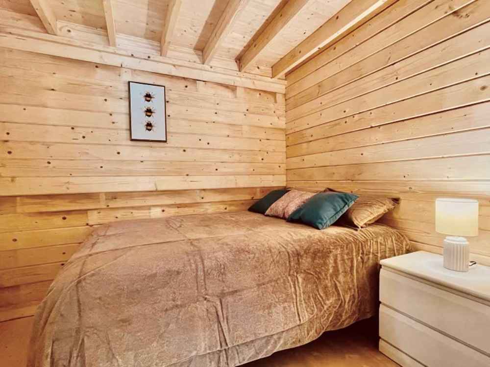 Snug bedroom in the Sawmill Structures Edwin Lake House PLUS 70MM Garden Room features wood-paneled walls, a slanted ceiling, a soft beige bedspread, and a small nightstand with a lamp