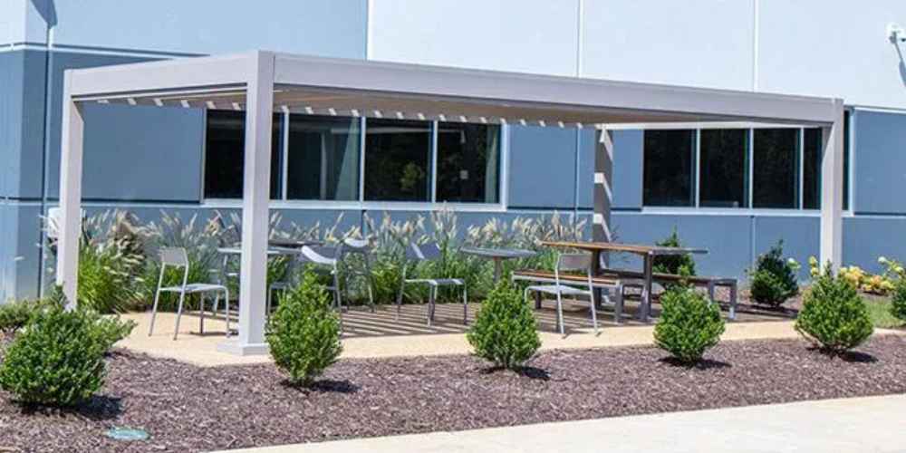 Structureworks Stratus Aluminum Pergola shelters a casual outdoor dining area with metal chairs and tables in front of a glass-windowed building