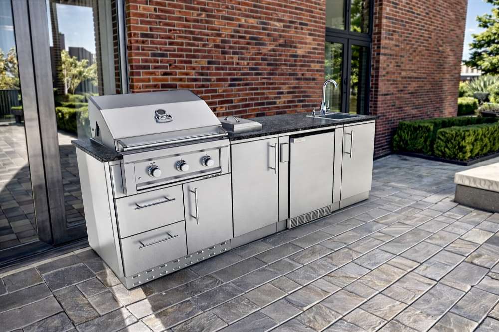 Sunstone Bayou 8ft 6in Outdoor Kitchen Island Package beside a brick home with grill, sink, and cabinet storage