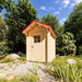 The Wee Wee House 33 with its windowed wooden door closed, red gabled roof and pale wood exterior surrounded by rocks, grasses and dense green trees.