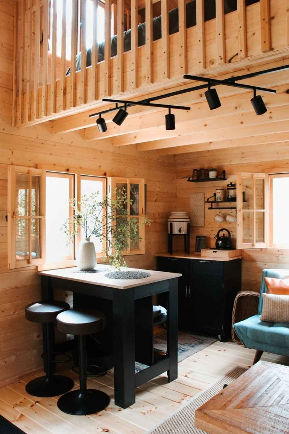 The dining and kitchen area of the Sawmill Structures BlueWater Loft 160 38mm Garden Room includes a black dining table, barstools, and open shelves with mugs and plants