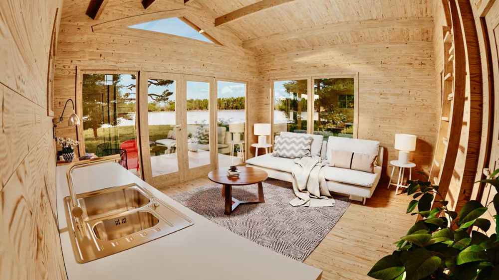 The kitchen sink and counter of the Sawmill Structures Edwin Lake House 70MM Garden Room face a bright and airy living room with large windows and comfy seating