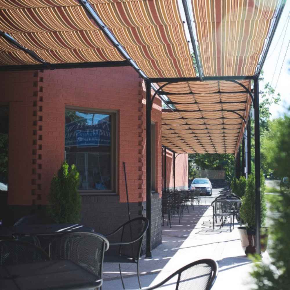 The long sidewalk patio of a restaurant is lined with tables beneath a colorful striped Structureworks Silhouette Aluminum Patio Cover & Pergola