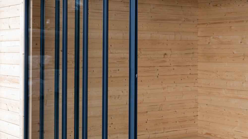 The tall, vertical glass panels of the GardenHouse24 USA ALU Concept 44Q Garden Room are neatly aligned in a wooden wall