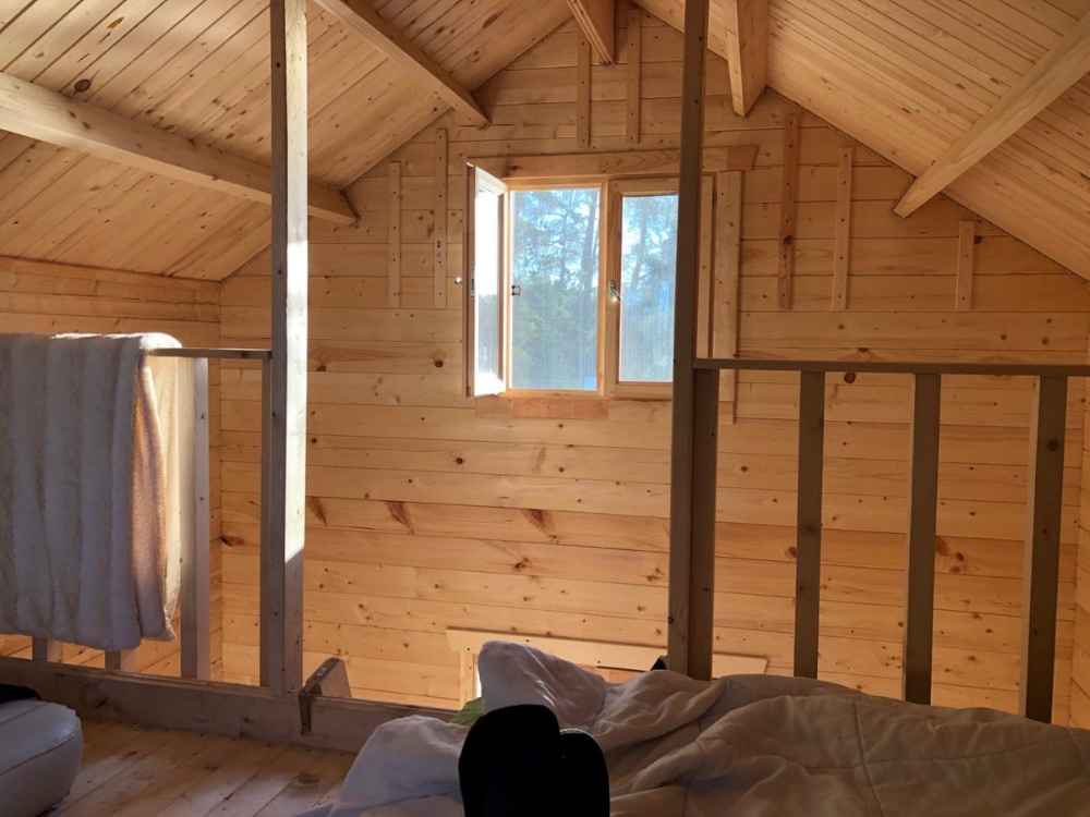 The upstairs loft of the Sawmill Structures Grand Loft 107 Deluxe DD Garden Room shows warm sunlight streaming through a small open window, casting light on wooden walls and a bed