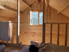 The upstairs loft of the Sawmill Structures Grand Loft 107 Deluxe DD Garden Room shows warm sunlight streaming through a small open window, casting light on wooden walls and a bed