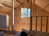 The upstairs loft of the Sawmill Structures Grand Loft 107 Deluxe DD Garden Room shows warm sunlight streaming through a small open window, casting light on wooden walls and a bed