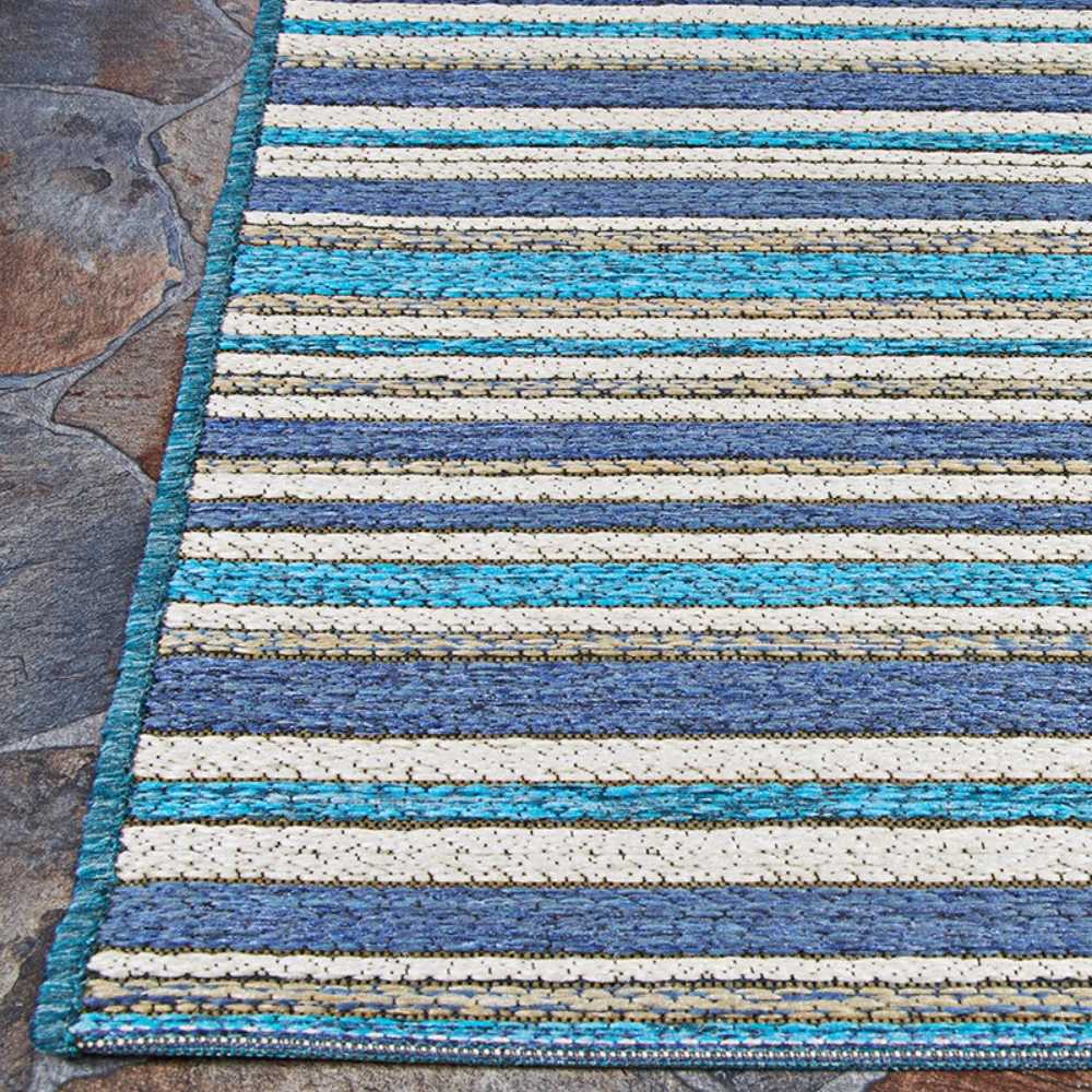 The corner of the Couristan Cape Indoor/Outdoor Rug - Brockton/Cobalt-Teal is shown on a stone patio, highlighting its bold horizontal stripe pattern and clean edge finish