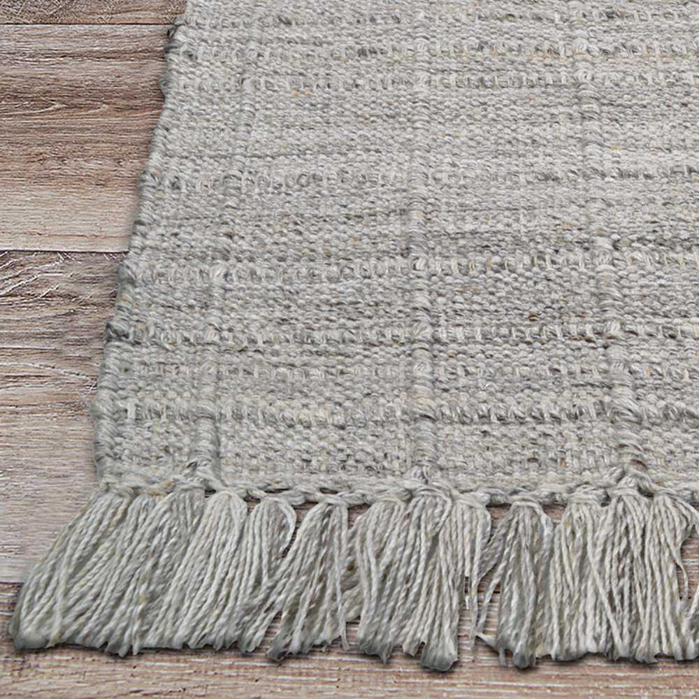 The edge of the Couristan Ascot Indoor/Outdoor Rug - Windowpane/Linen lies flat on a wooden floor, featuring thick tassels and a woven pattern