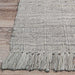 The edge of the Couristan Ascot Indoor/Outdoor Rug - Windowpane/Linen lies flat on a wooden floor, featuring thick tassels and a woven pattern
