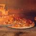 The image captures a pizza in a brick oven being adjusted with the Chicago Brick Oven Aluminum 17.5 Inch Pizza Spinner Turning Fork (2-Pack) during cooking