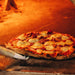 The image displays a hot pizza being turned inside a wood-fired oven using the Chicago Brick Oven Aluminum 17.5 Inch Pizza Spinner Turning Fork (2-Pack)