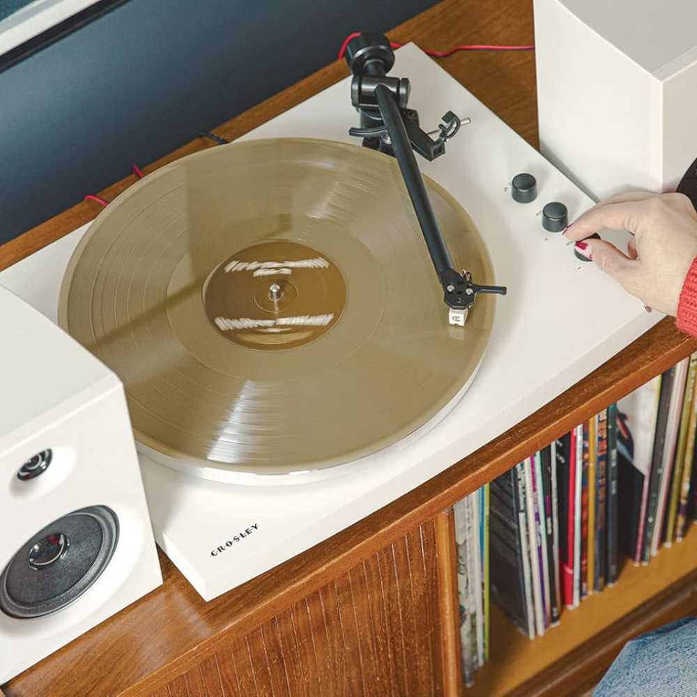 The image shows a Crosley T150 T-Series Turntable System - White with a spinning gold vinyl record, a person adjusting the volume knob, and a white speaker on a wooden shelf filled with vinyl