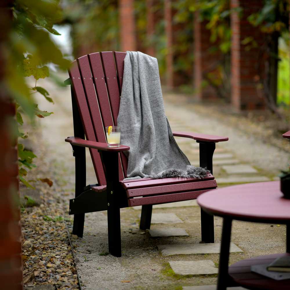 The image shows a cherrywood and blackLuxCraft Royal Adirondack Chair outdoors on a stone pathway, with a gray blanket draped over the back and a drink resting on the armrest