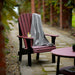 The image shows a cherrywood and blackLuxCraft Royal Adirondack Chair outdoors on a stone pathway, with a gray blanket draped over the back and a drink resting on the armrest