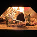 The image shows a fire blazing around a metal structure inside a brick oven, featuring the Chicago Brick Oven Steel Log Holder supporting the burning logs