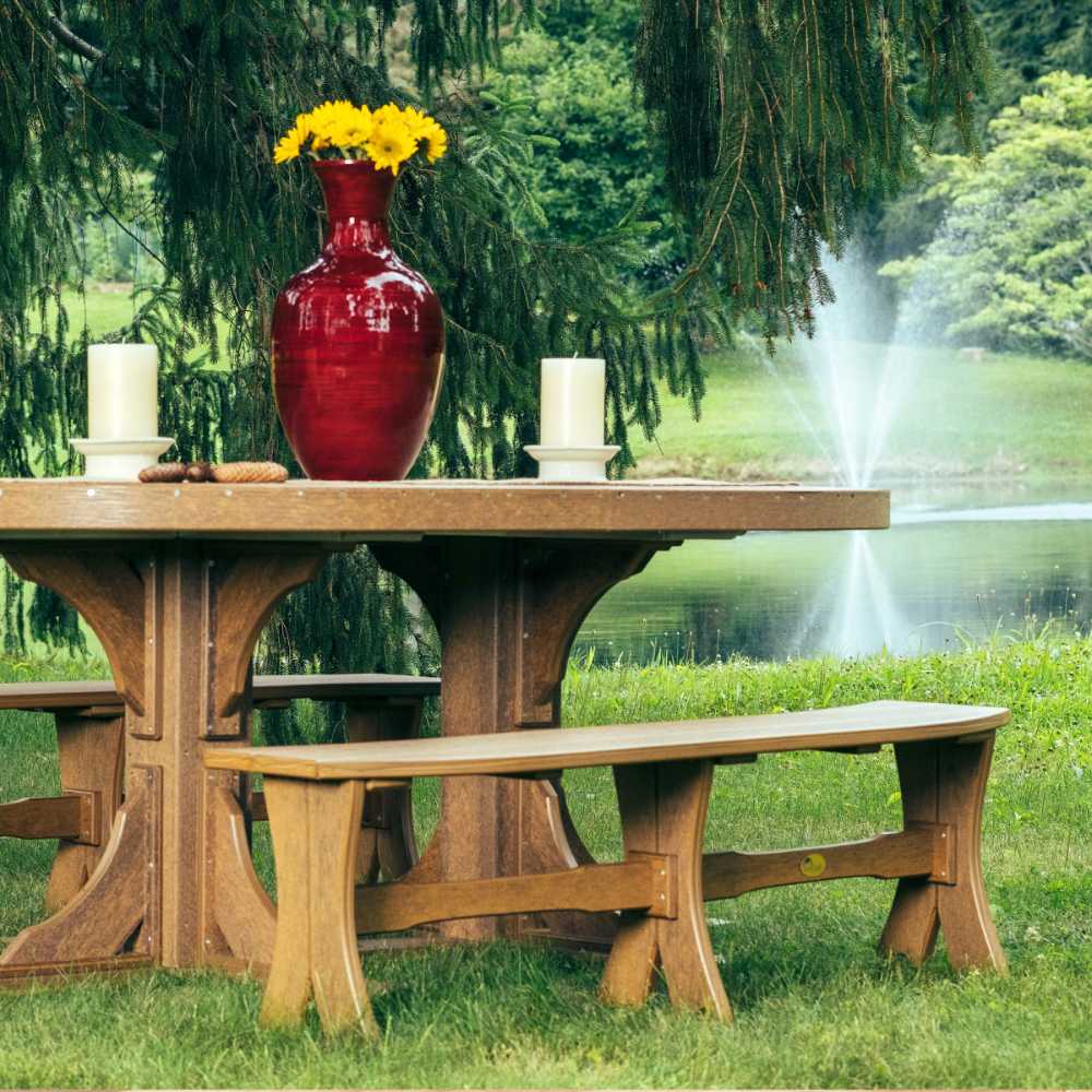 The image shows an outdoor scene with LuxCraft 52 Table Bench on green grass by a lake with a fountain in the background, a large red vase with sunflowers, and candles on the table