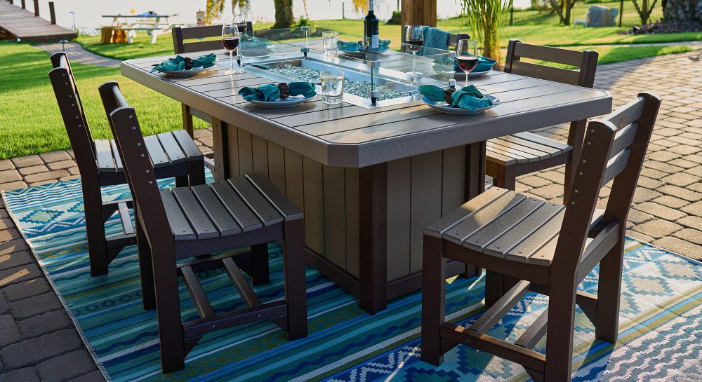 The image shows a square fire table with a glass-protected fire pit surrounded by matching slatted wooden chairs, part of the LuxCraft Luminary Furniture Set, on a colorful outdoor rug