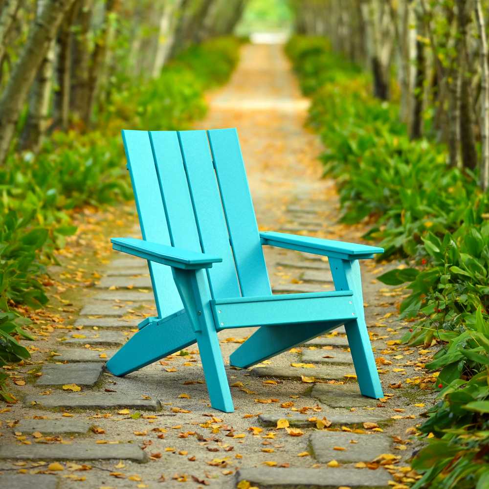 The image shows a vibrant aruba blue LuxCraft Urban Adirondack Chair in the middle of a garden path surrounded by green bushes and autumn leaves